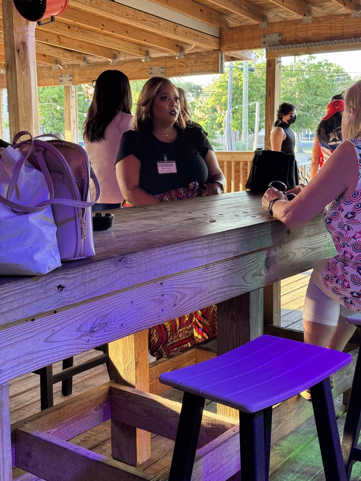 A woman wearing a name tag stands at a wooden bar outdoors during the Hispanic Heritage Month kick-off. Sunlight streams in as several people, bags and stools around them, engage in lively conversation—setting the scene for Sept 2025 festivities.