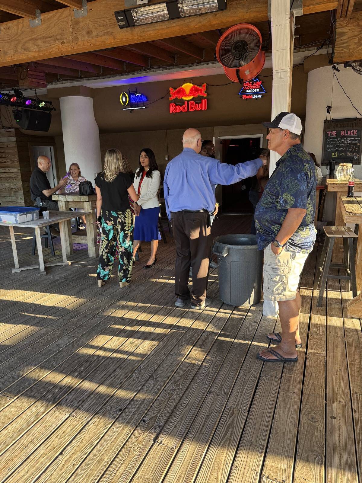 A group of people socialize on a wooden deck at a bar with neon signs on the wall during a vibrant 2025 Hispanic Heritage Month kick-off. One man stands near a trash can, while others chat in small groups as sunlight casts shadows across the deck.