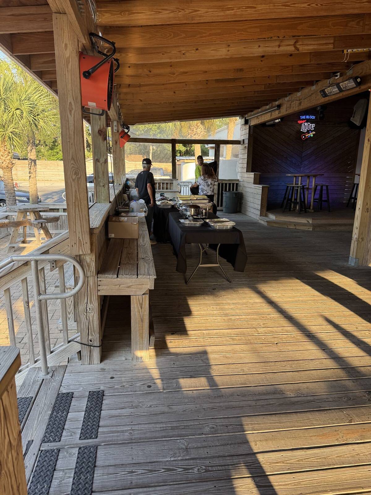 A wooden outdoor patio with benches and tables, where two people stand behind a black table set up with buffet trays for a Kick-off to Hispanic Heritage Month. Sunlight streams in, palm trees are outside, plus empty bar stools and a neon sign.