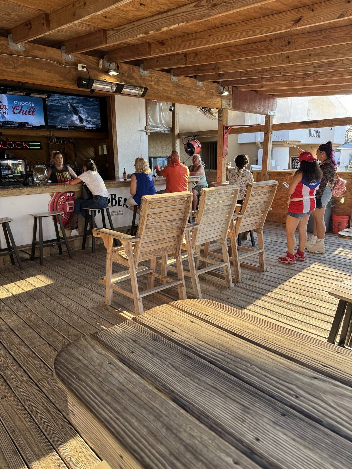People sit and stand at a wooden outdoor bar with high stools under a wooden roof. As the bartender serves drinks and TVs are mounted above, sunlight streams in, creating a relaxed vibe—perfect for a Hispanic Heritage Month kick-off gathering.