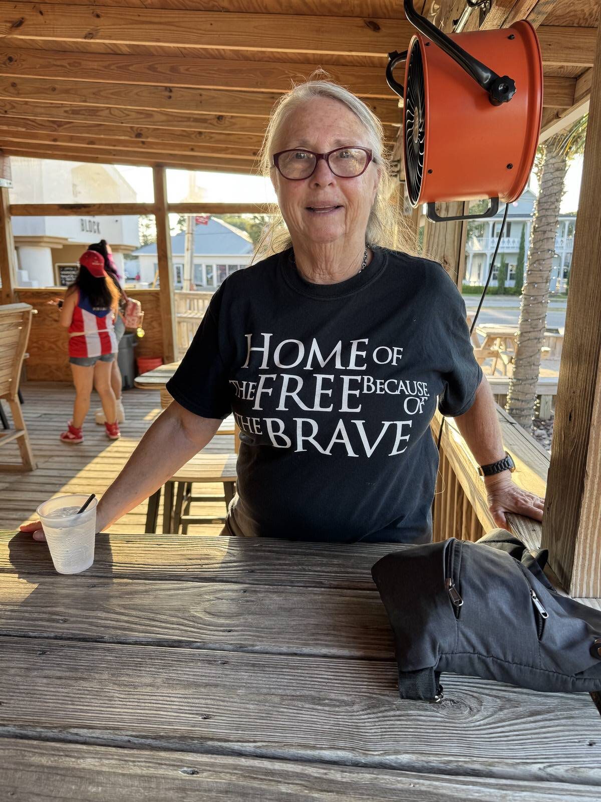 Older woman with long blond hair and glasses stands at a wooden bar, smiling, during the Hispanic Heritage Month Kick-off. She wears a black shirt reading "Home of the Free Because of the Brave." A black bag and drink sit on the bar; a fan is above her.