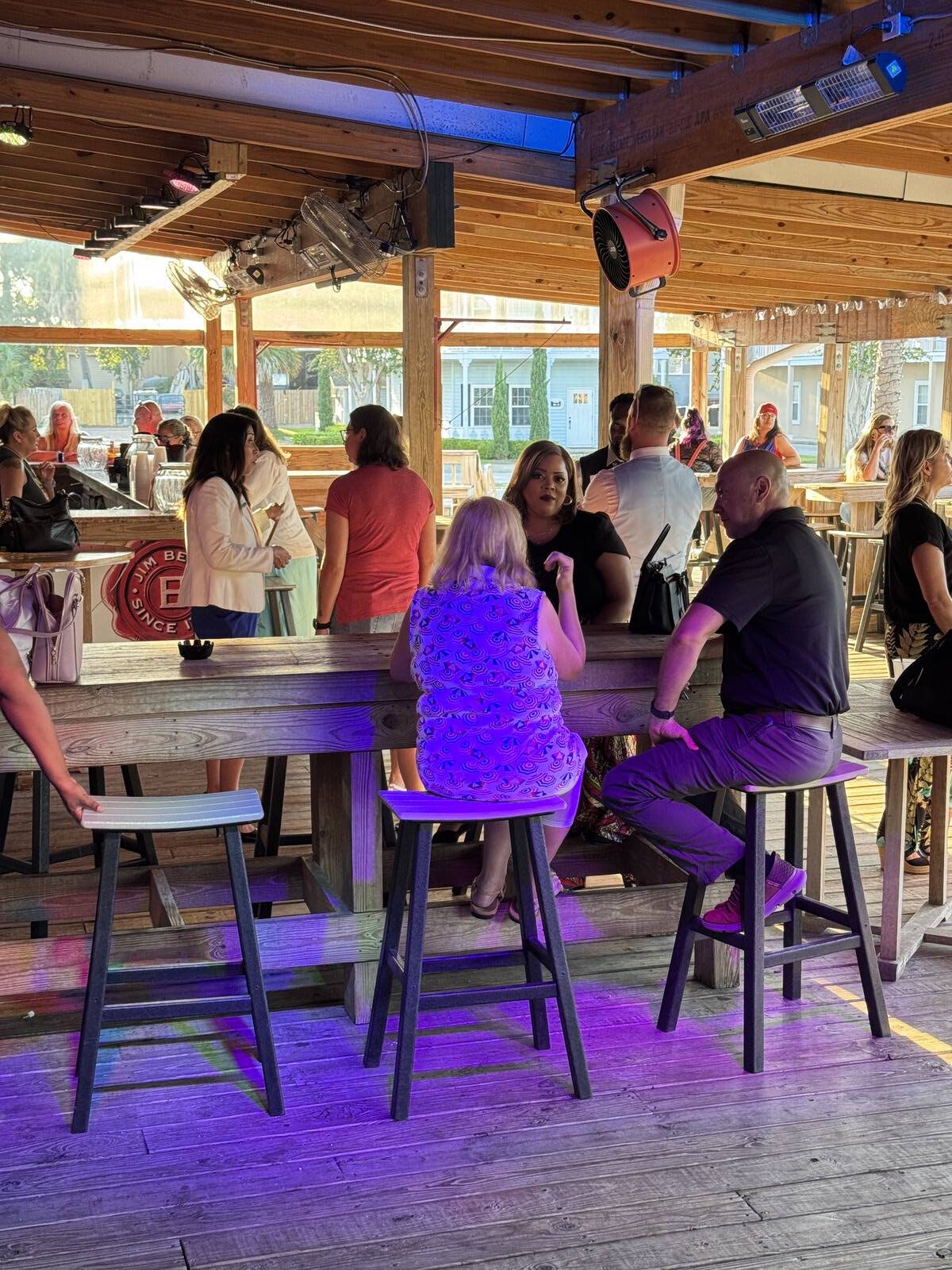 People are sitting and standing around wooden tables at an outdoor bar with a wooden roof, enjoying drinks and colorful lights on the floor, creating a lively social atmosphere perfect for a Hispanic Heritage Month Kick-off in Sept 2025.