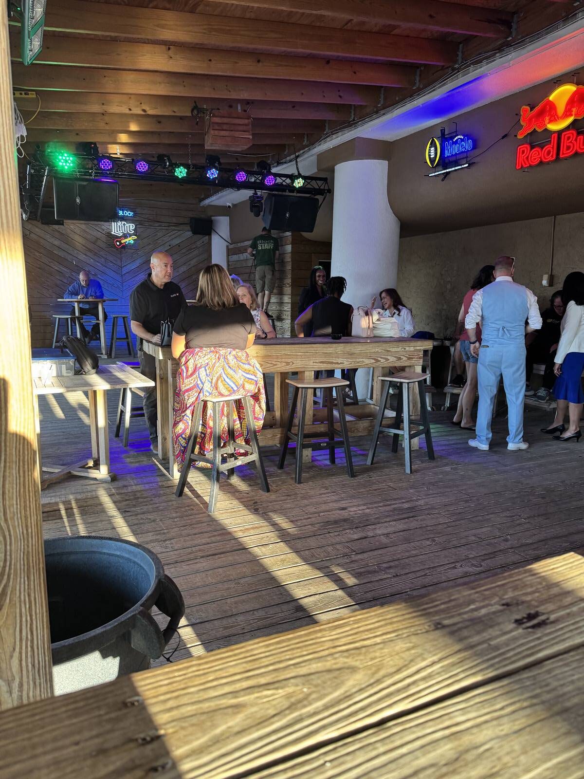 People sit and stand around wooden tables in a lively indoor-outdoor bar with wooden floors and ceiling, celebrating the kick-off of Hispanic Heritage Month. Colorful lights and neon drink signs add to the festive September 2025 atmosphere.