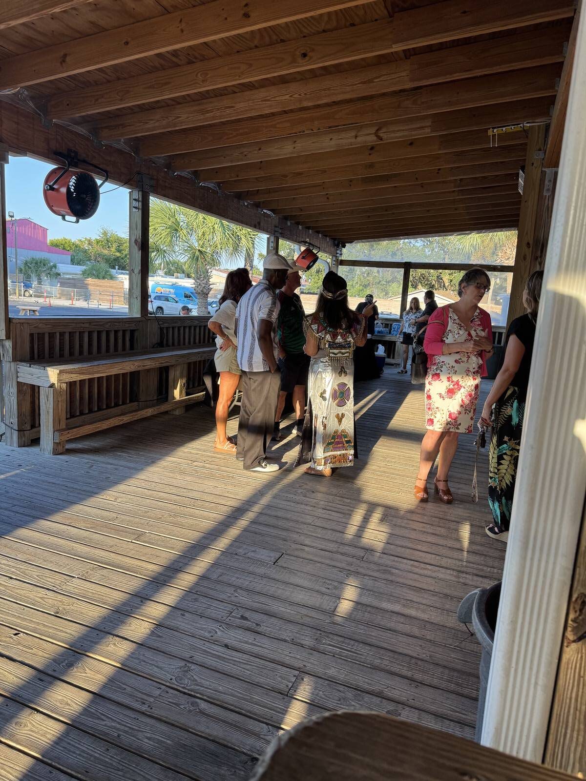 A group of people stands and talks on a wooden covered deck in the sunlight during a Hispanic Heritage Month kick-off. Some wear floral dresses, others are in casual attire. Palm trees and parked cars are visible outside.