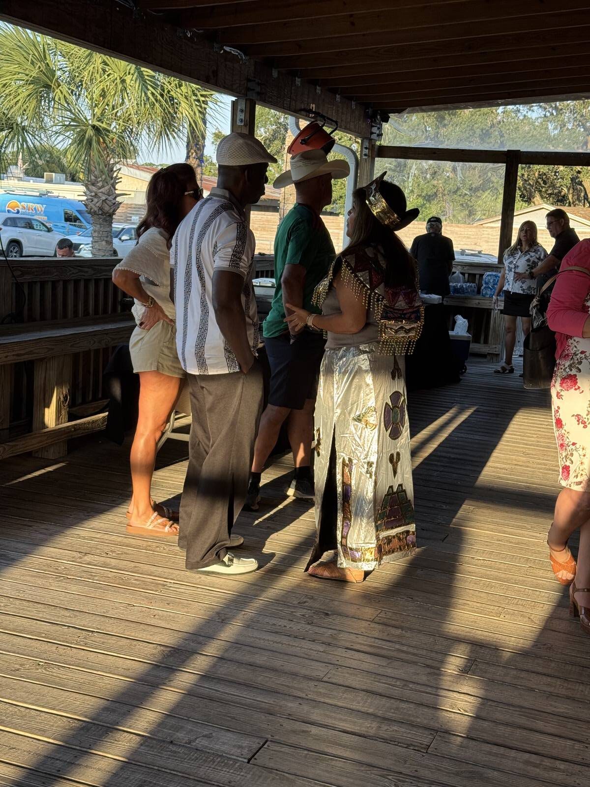 People in costume, including a woman dressed as Cleopatra and two men in hats, gather and talk on a wooden deck during the Hispanic Heritage Month Kick-off. Others stand near tables by the wooden railing, with trees and cars visible outside.