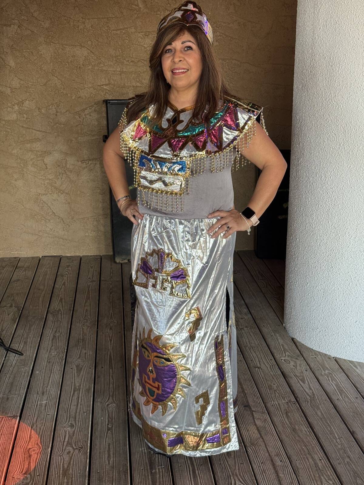 A woman stands on a wooden deck wearing a shiny, colorful costume with gold, purple, and turquoise details, geometric patterns, and a decorative headdress, smiling at the camera at the 2025 Hispanic Heritage Month Kick-off.