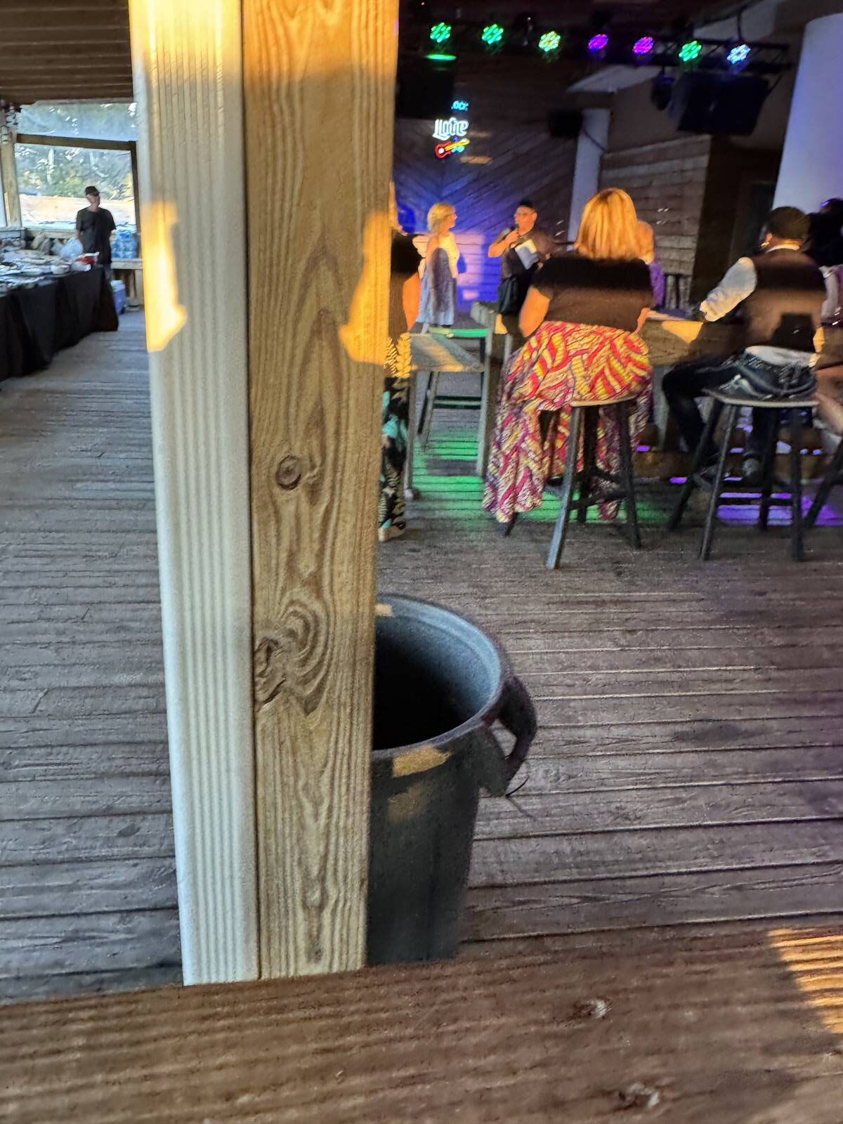 A wooden post divides the image, with a trash can at its base. In the background, people sit at tall tables on a wooden deck, enjoying a Hispanic Heritage Month Kick-off under colorful lights in this casual indoor-outdoor setting.