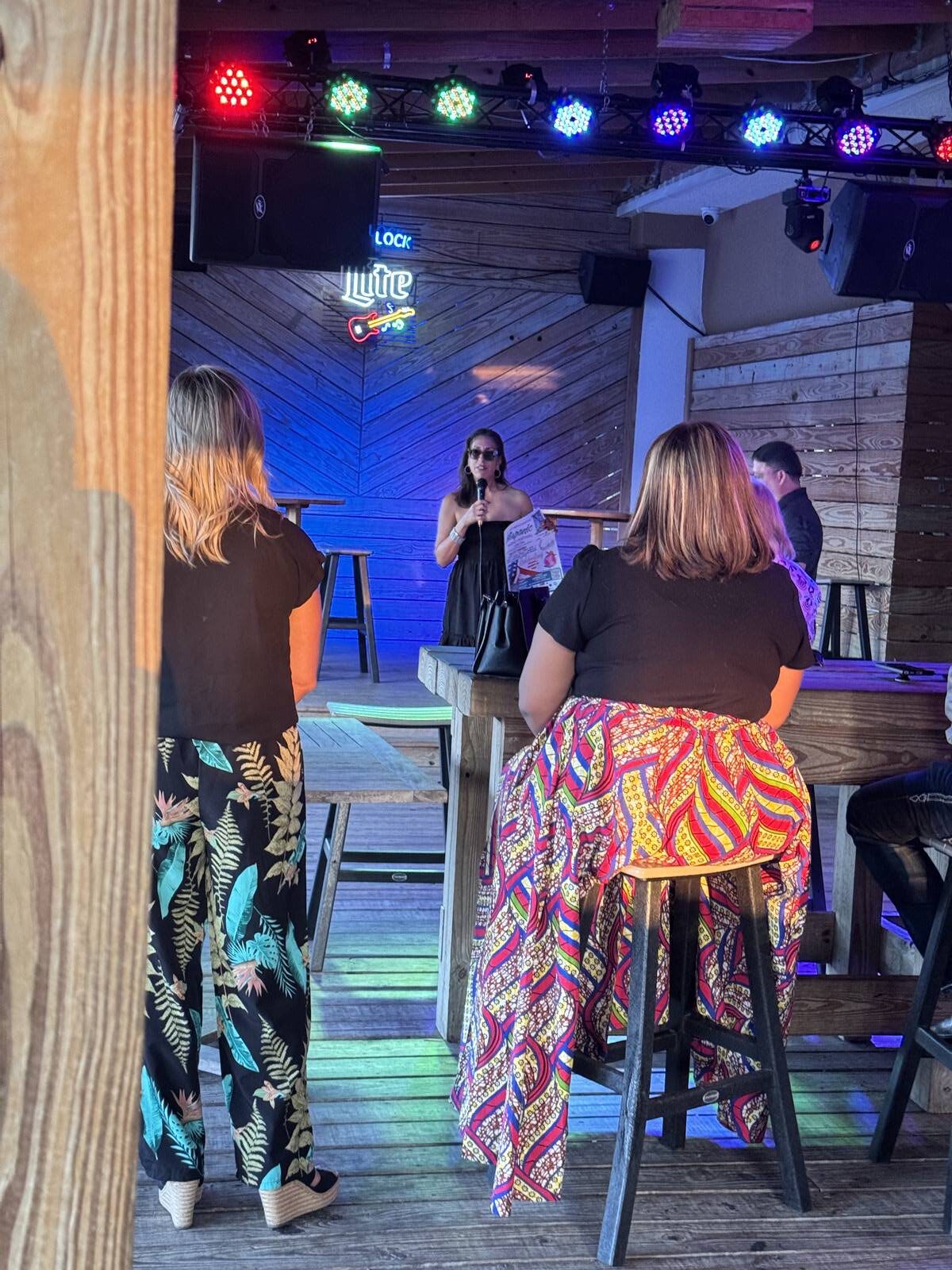 A woman sings on stage in a wooden bar with colorful lights above her, kicking off Hispanic Heritage Month celebrations. Two women with patterned pants sit at a table in the foreground, enjoying the relaxed and casual atmosphere.