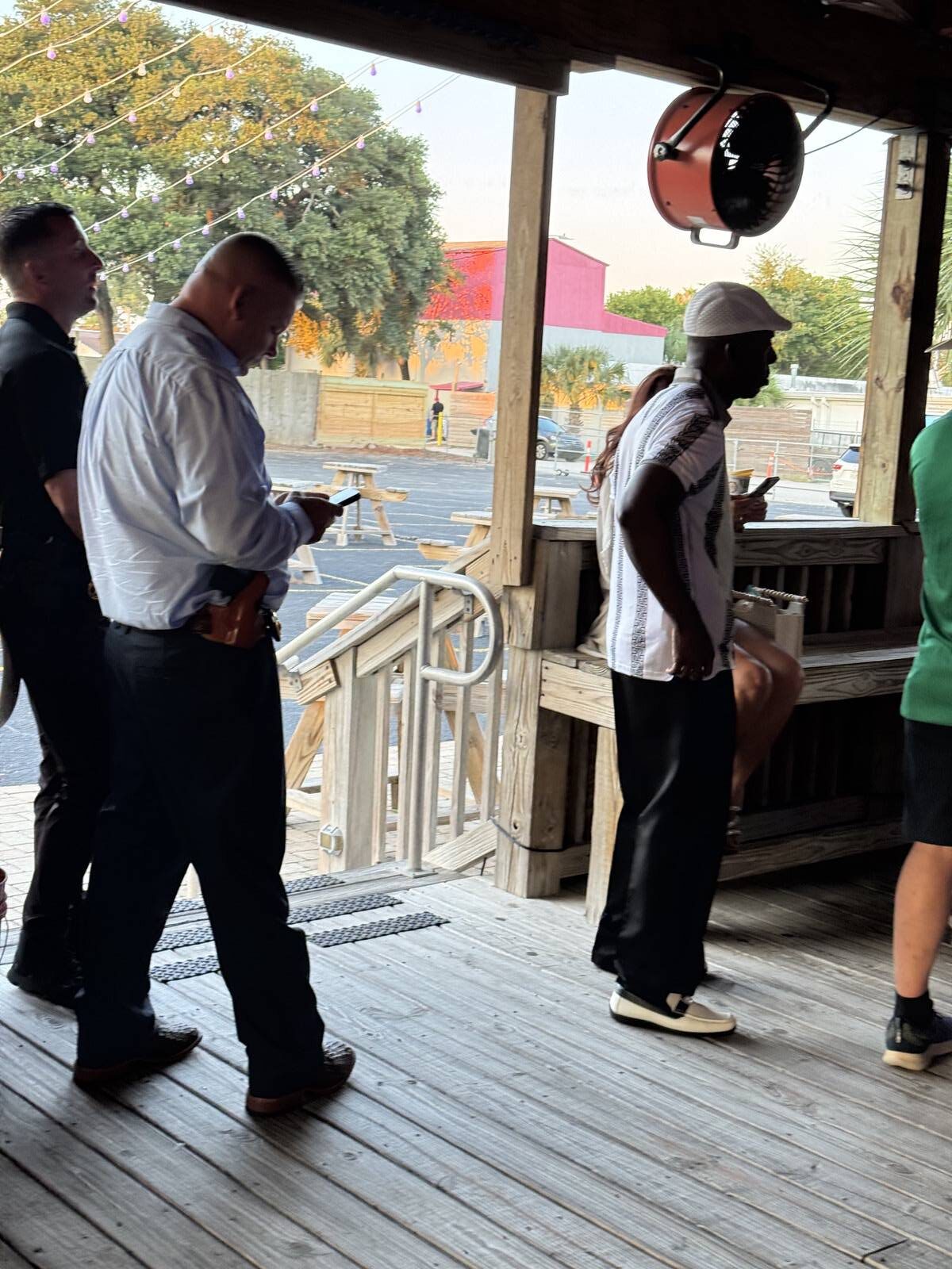 Three men stand in line on a wooden deck outside—one with a phone and gun holster, another in a white hard hat, and the third in black—during the kick-off for Hispanic Heritage Month. Picnic tables and trees are visible in the background.