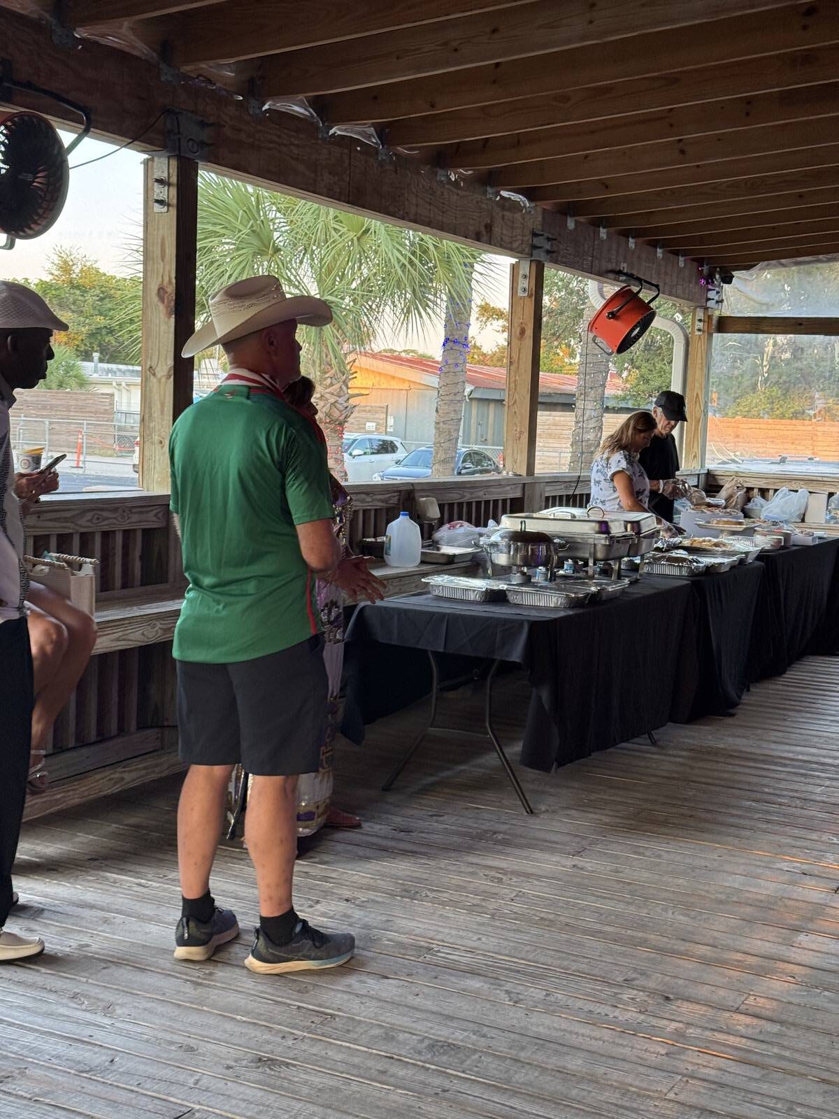 People are standing in line at a buffet table under a wooden pavilion during the Sept 2025 Kick-off for Hispanic Heritage Month. Food trays and drinks are set out, a palm tree is visible outside, and the atmosphere appears casual and relaxed.