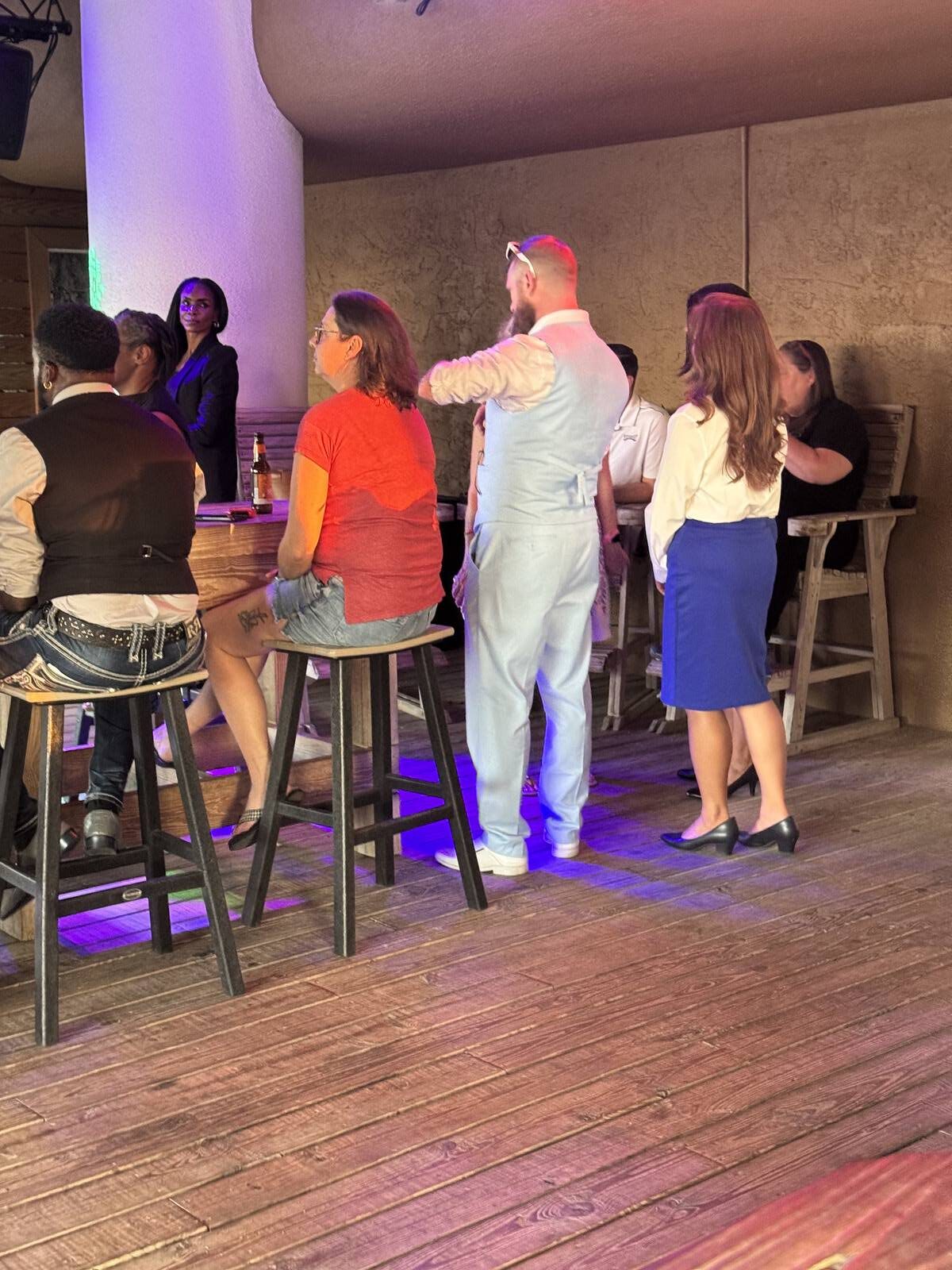 A group of people sit and stand around a wooden bar indoors during a Sept 2025 Kick-off. Two standing in business attire, others seated in casual clothes, as purple and blue lighting reflects on the floor—perfect for Hispanic Heritage Month celebrations.