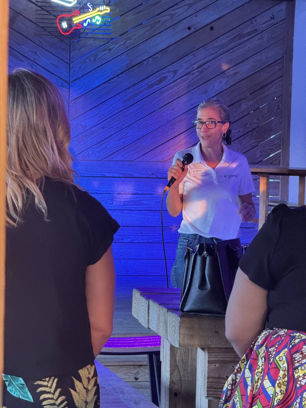 A woman in a white shirt and glasses speaks into a microphone on a small stage, holding papers. Two people stand in the foreground, listening. The setting has wooden walls and blue lighting, celebrating Hispanic Heritage Month.