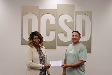 Two people stand in front of large letters spelling "OCSD" on a wall. The woman on the left is handing an envelope to the man on the right, celebrating a community partnership to support students. Both are smiling.