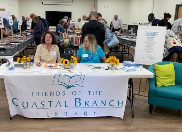 Two women sit at a table with a “Friends of the Coastal Branch Library” banner, surrounded by sunflowers, as people browse tables filled with books at the busy Coastal Branch Library book sale event.