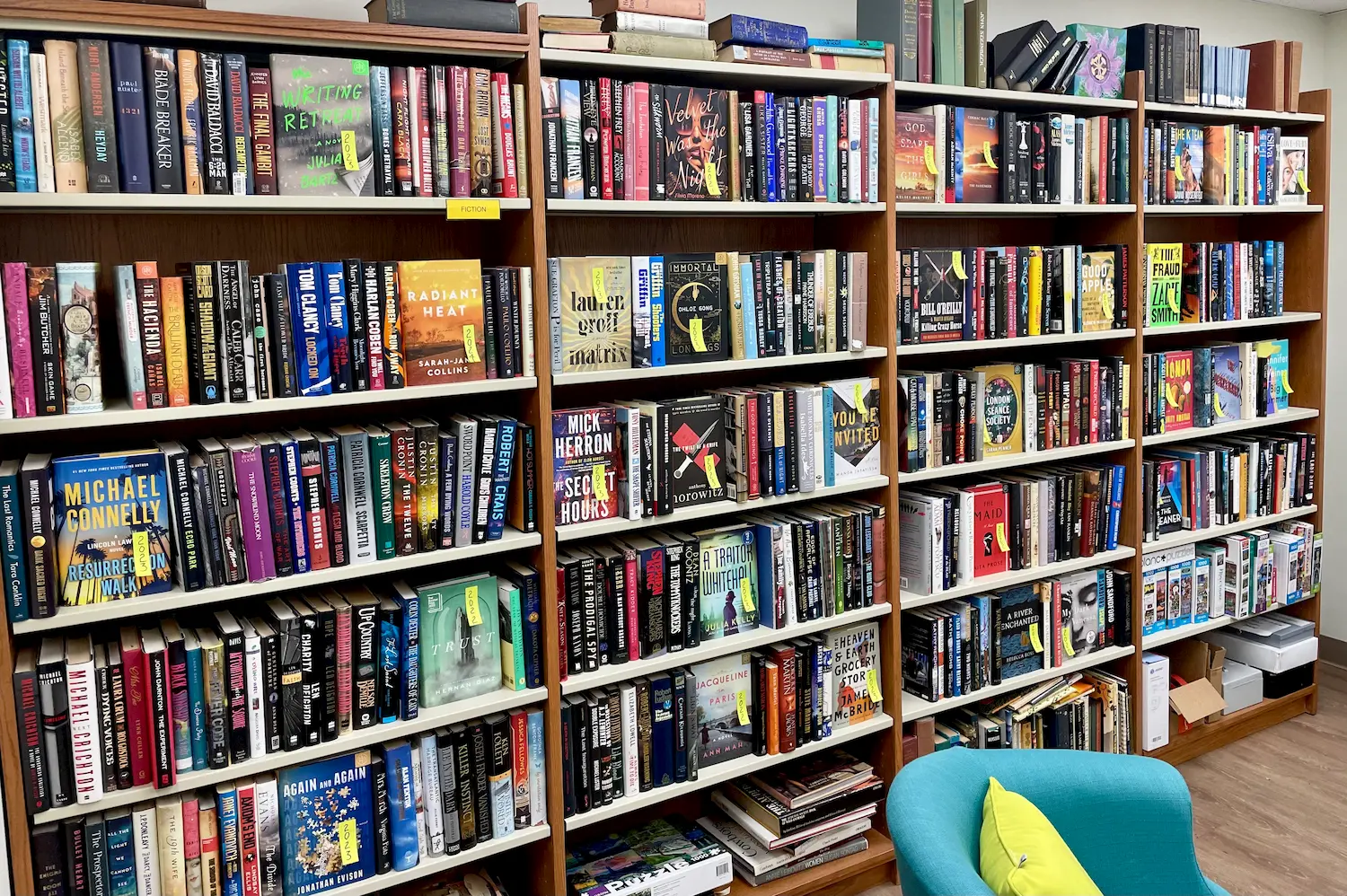 Bookshelves filled with colorful books in the Coastal Branch Library, with a light blue chair in the foreground. Friends of the Library boxes and binders are on the bottom shelves to the right, hinting at an upcoming book sale.