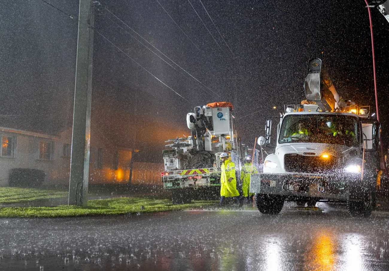 Utility workers in rain gear repair power lines at night during heavy rain, using large utility trucks with bright lights on a residential street.