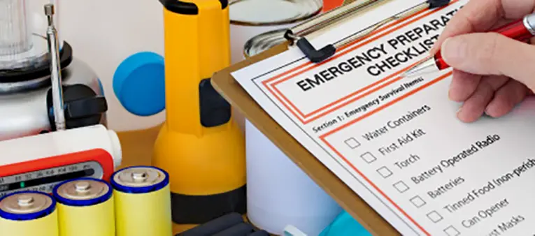 A hand holds a pen over an emergency preparedness checklist on a clipboard. Surrounding items include batteries, a flashlight, a radio, a first aid kit, and other emergency supplies.