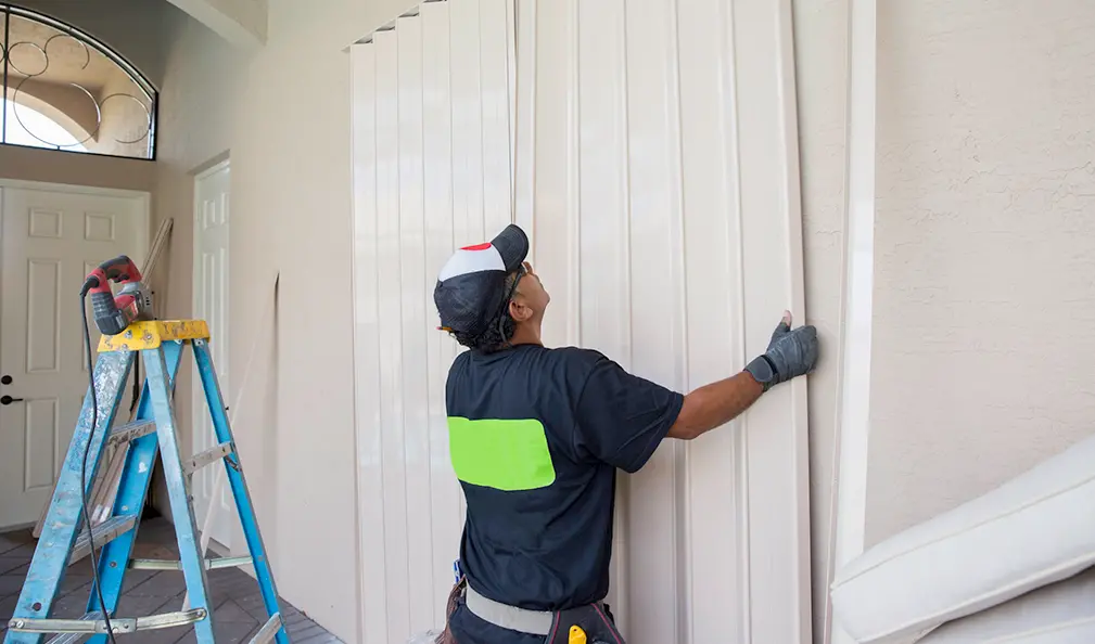 A person in work gloves installs white hurricane shutters on a house exterior near a blue stepladder and an open front door.