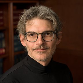 A middle-aged man with gray hair, glasses, and facial hair wears a black turtleneck. He is posed in front of shelves filled with books, looking directly at the camera—an image celebrating Hispanic Heritage Month.