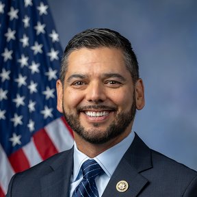 A man in a dark suit, white shirt, and striped tie smiles in front of an American flag and a blue backdrop, proudly wearing a circular pin on his lapel as he celebrates Hispanic Heritage Month. He has short dark hair and a beard.