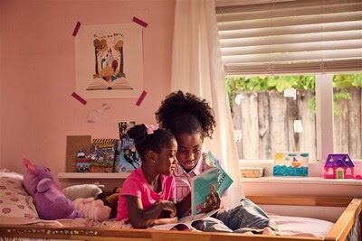 Two young girls sit together on a bed in a cozy, sunlit bedroom, reading a book. Surrounded by plush toys and colorful drawings, they discover a love with books—just like many Florida students in the free program. Sunlight streams through window blinds.