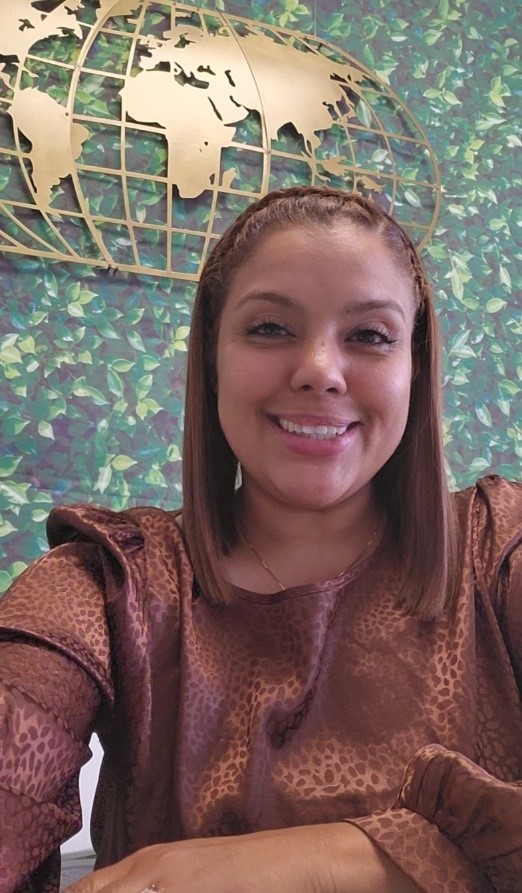 A woman with straight, shoulder-length hair smiles at the camera. She is wearing a brown patterned blouse and sits in front of a green leafy wall, adorned with a gold world map and subtle palm tree accents.