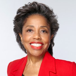 A smiling woman with short dark hair, wearing a bright red blazer and red lipstick, poses in front of a plain light background, celebrating Herencia Hispana during Hispanic Heritage Month.