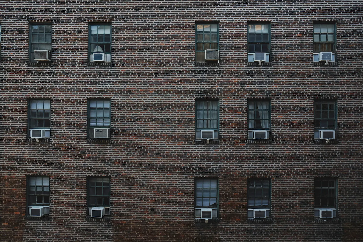 A brick apartment building facade with multiple rows of identical windows, each window equipped with a window-mounted air conditioning unit—an all-too-common scene as rising energy costs drive residents to seek relief from the Florida energy bills.