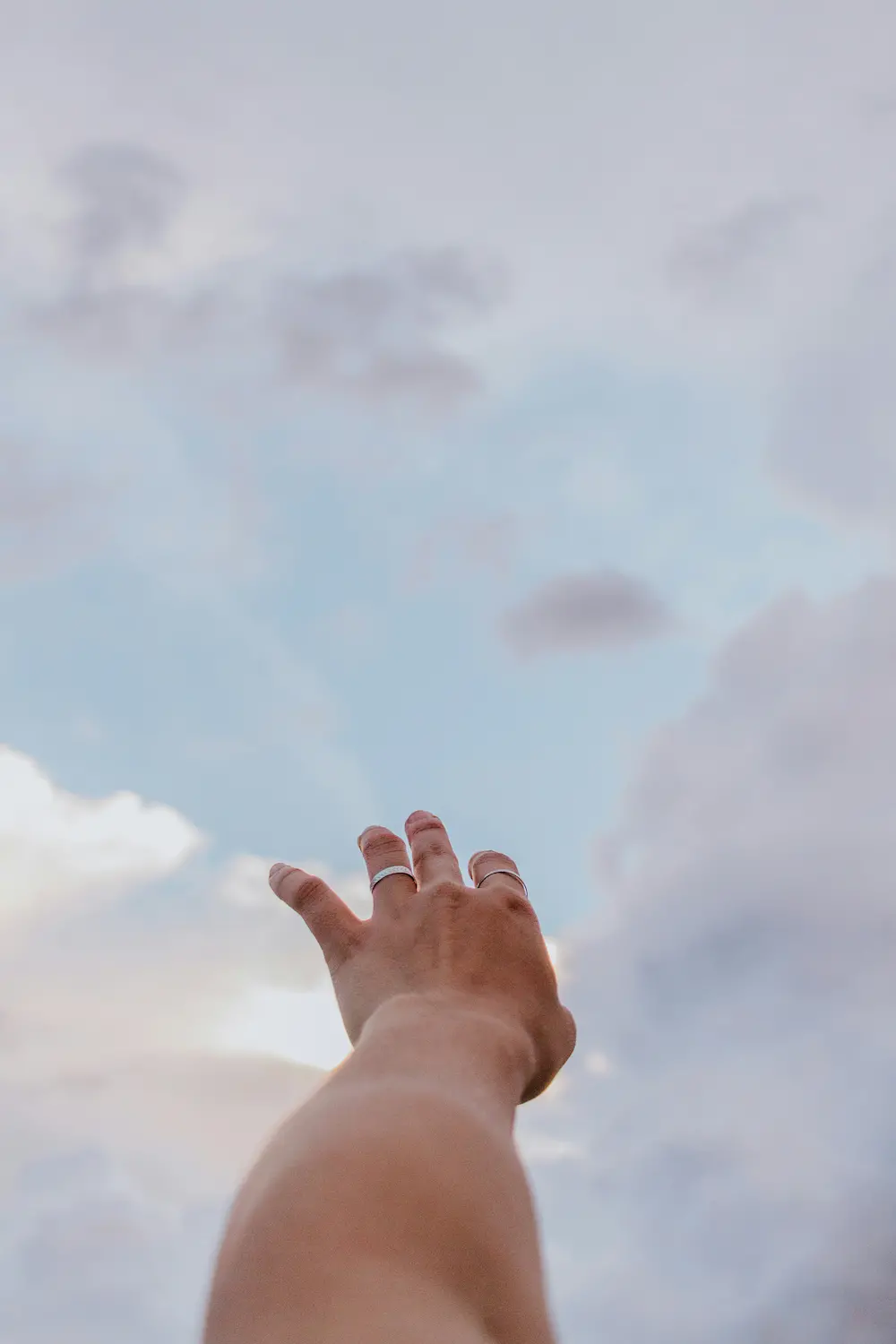 A person's outstretched hand reaches up toward a cloudy sky, as if reaching for heaven, with soft light illuminating the scene and blue and gray clouds in the background.