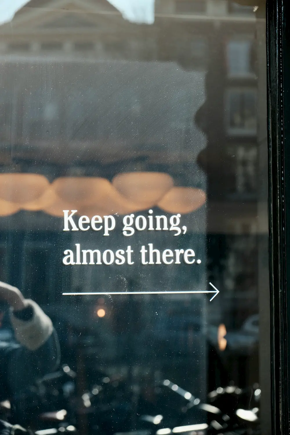 White text on a glass window reads, "Keep going, almost there," with an arrow pointing right—a motivating sight for new entrepreneurs starting a business. Blurry buildings and lights are reflected in the window, adding to the inspirational scene.