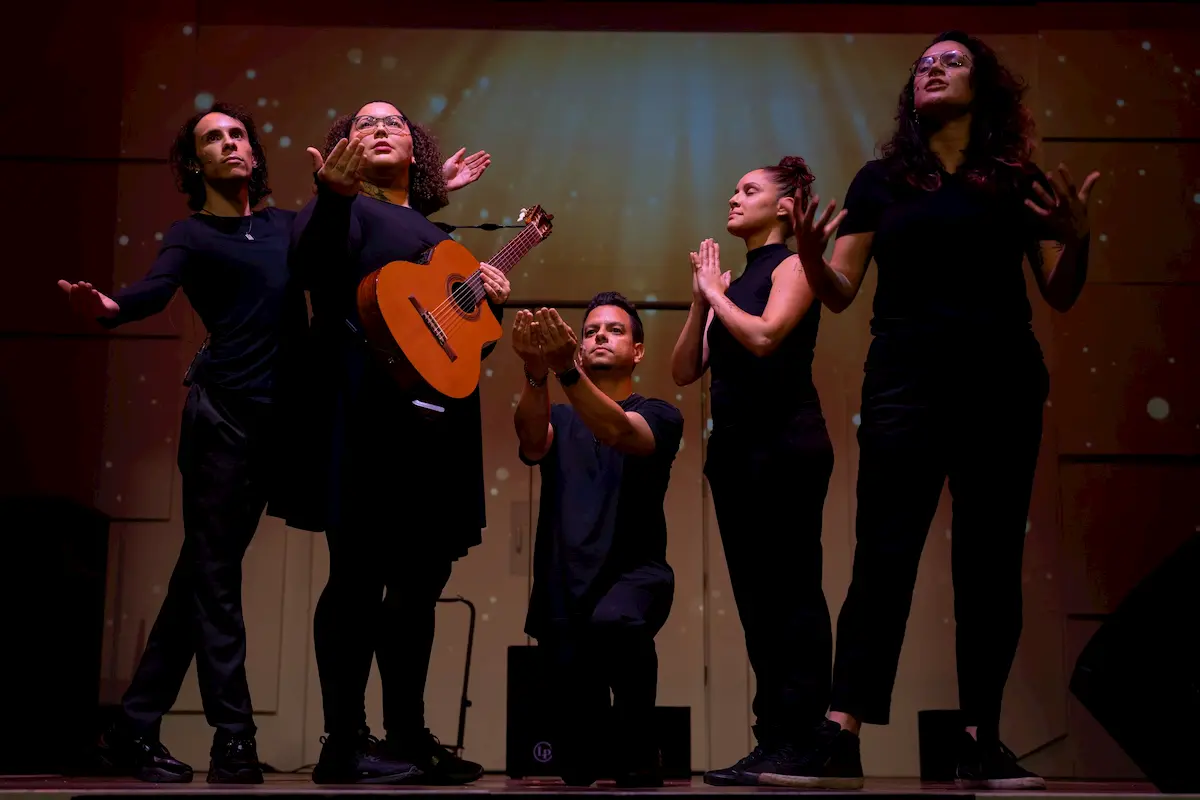 Five people on stage dressed in black perform dramatically under warm spotlights; guided by Descolonizarte TEATRO, they embody healing through art, with one holding a guitar while others strike expressive, community-building poses.
