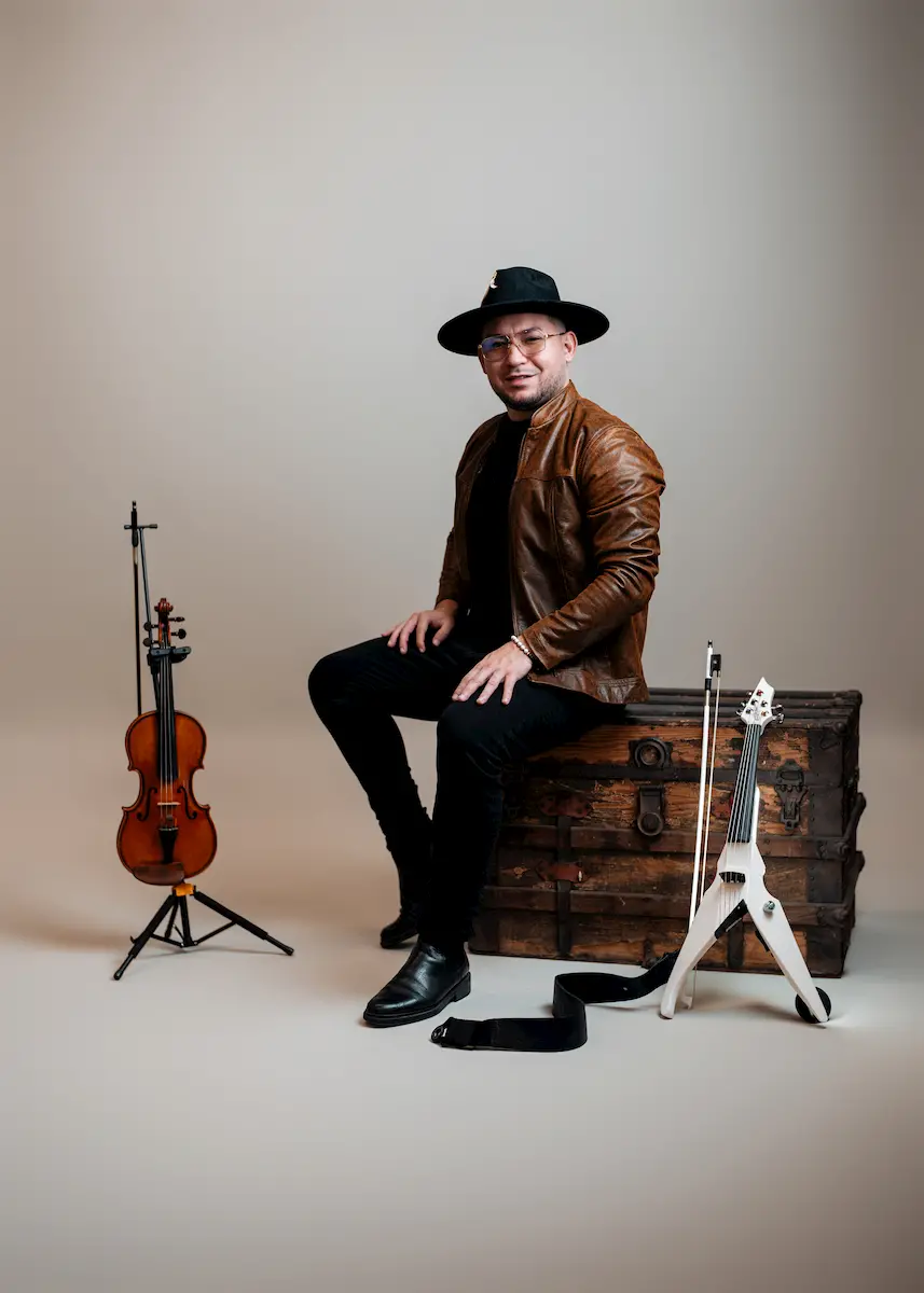 A man with international experience sits on a vintage wooden trunk, wearing a brown leather jacket and black hat. Beside him, a classical violin and a white electric violin hint at his journey as a violinist through South East music.