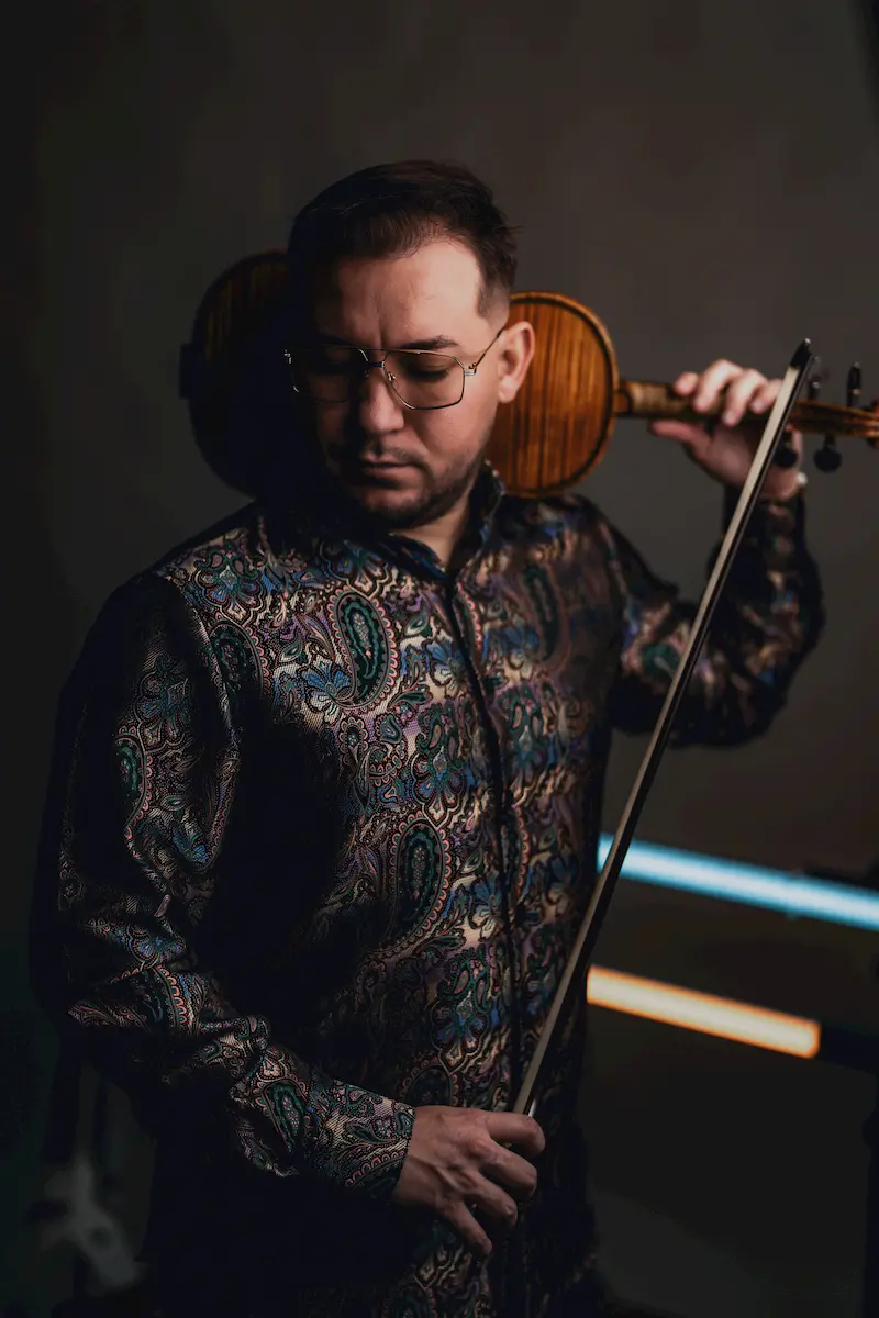 A violinist with international experience, wearing glasses and a patterned shirt, holds his violin on his shoulder and bow in hand, looking down with a serious expression in a dimly lit setting.