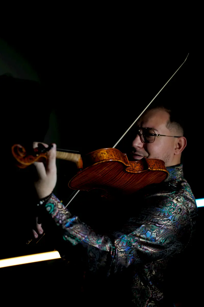 A violinist with international experience, wearing glasses and a colorful, patterned shirt, plays the violin with focus against a dark background, dramatic lighting highlighting the instrument and his face.