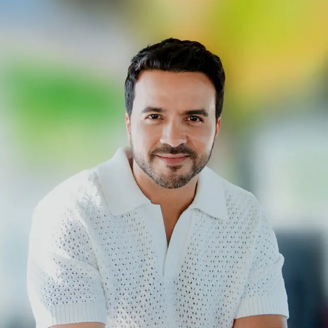 Un hombre sonriente, con pelo corto oscuro y barba, vestido con una camisa blanca de punto de manga corta, está sentado con los brazos cruzados sobre un fondo exterior difuminado con tonos verdes y amarillos.