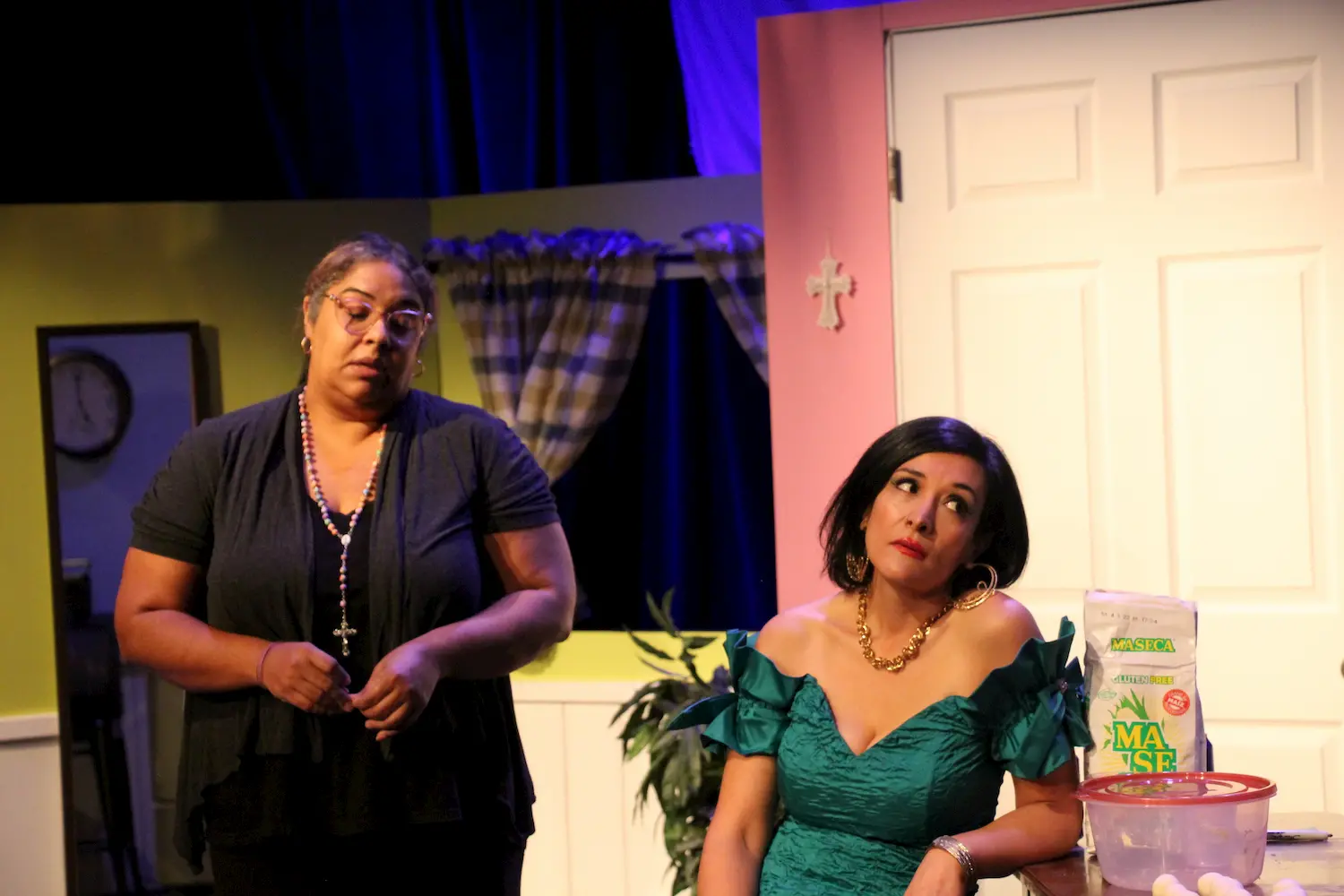 Two women on stage: one in glasses and black clothes holding a rosary looks serious, while the other in a green off-shoulder dress sits with an exasperated expression. They are in a colorful, domestic set with a white door and checked curtains.