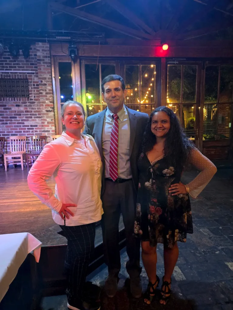 Three people smile and pose indoors at a dressed-up event; a man in a suit and two women, one in a floral dress and one in a white blouse, stand in front of brick walls and string lights.