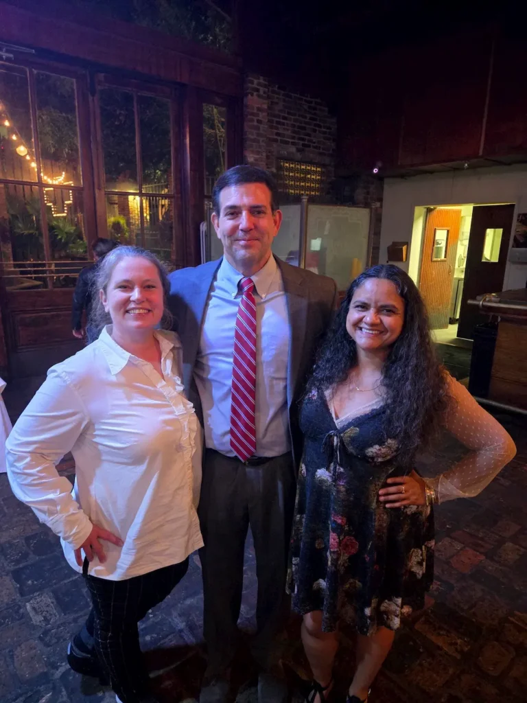 Three people pose and smile together indoors on a brick floor. The man in the middle wears a suit and red striped tie, while the two women on either side wear a white blouse and a dark floral dress. Warm lighting creates a cozy atmosphere.