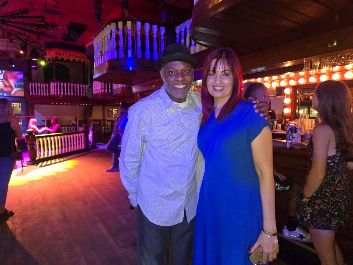 A smiling man in a hat and a woman in a blue dress pose together in a warmly lit bar or nightclub with wooden decor and people in the background.