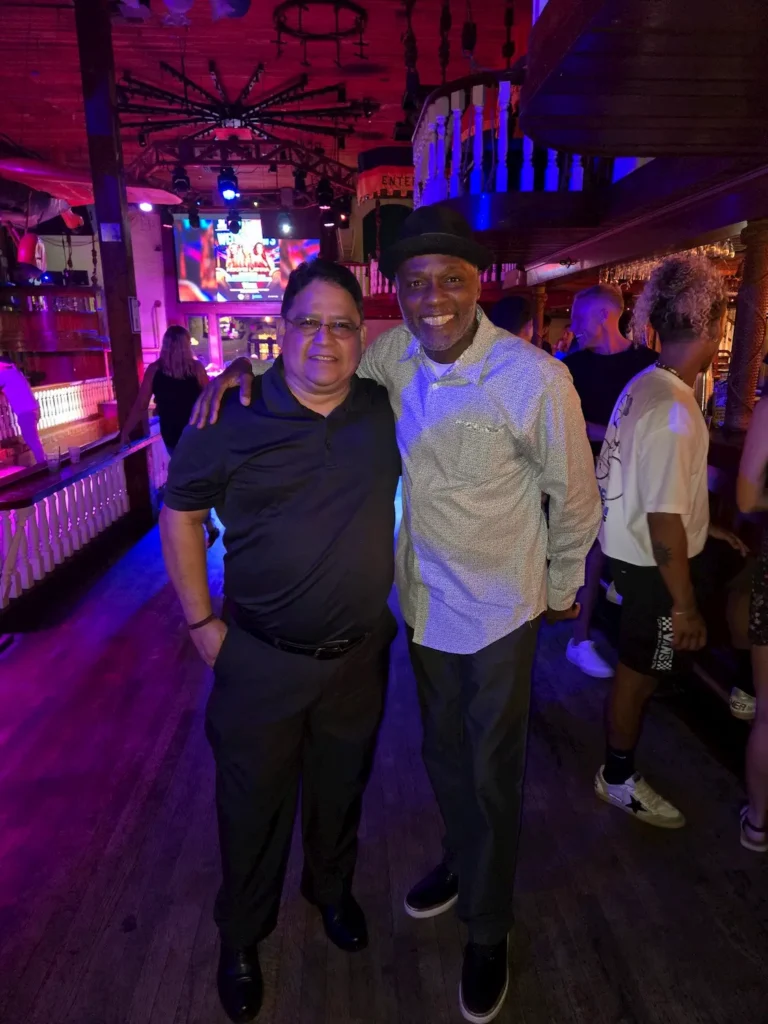 Two men pose and smile together inside a dimly lit bar or club with colorful lighting. Other people and bar decor are visible in the background.