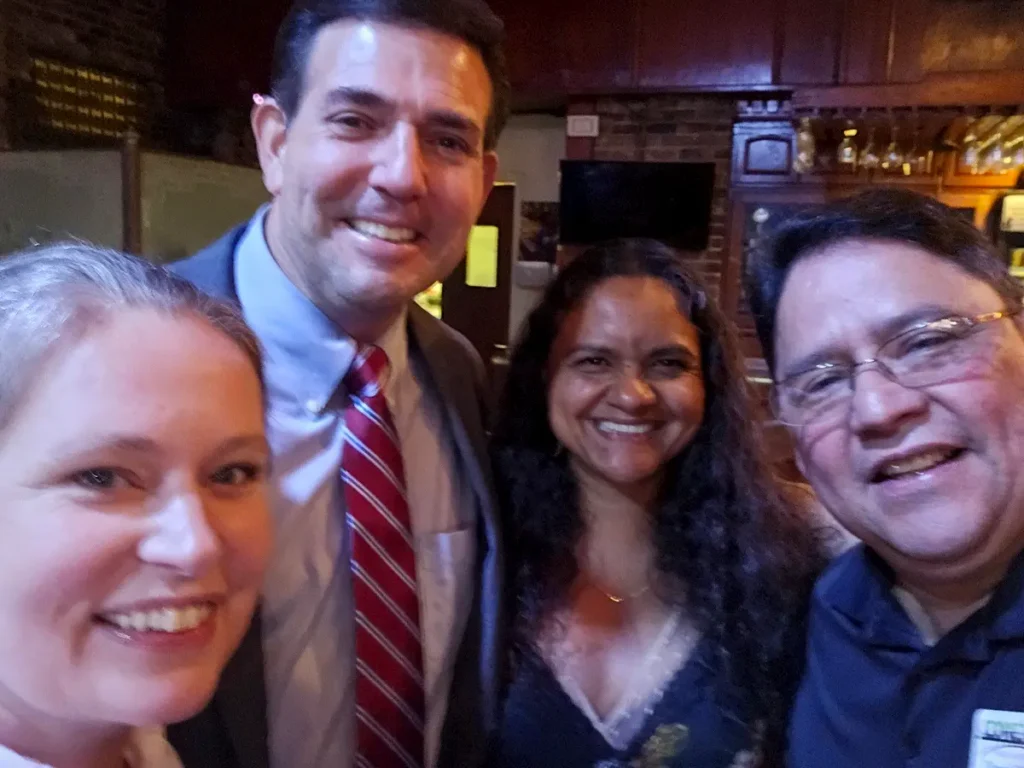 Four people smiling and posing together indoors, dressed in business or semi-formal attire, with a brick wall and framed pictures in the background.