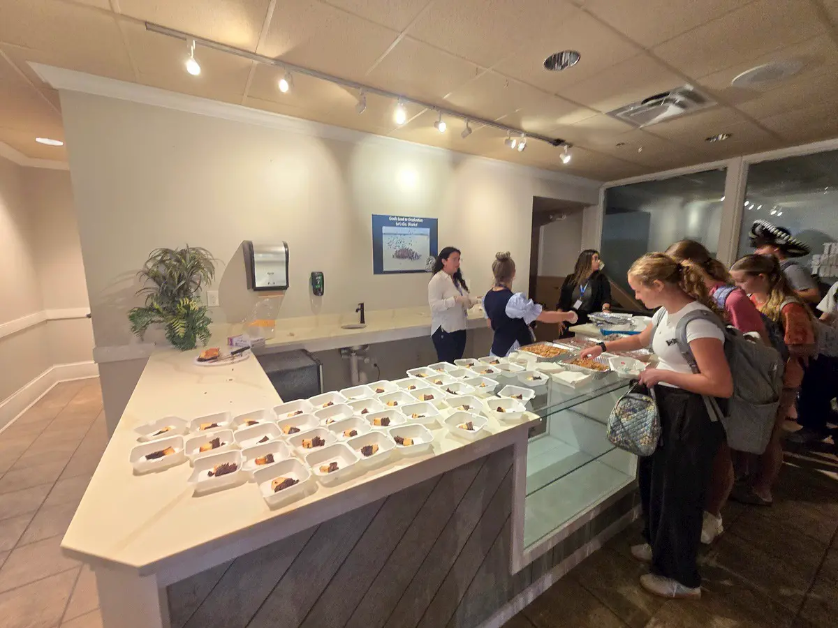 Students stand in line at a cafeteria counter with trays of food in takeout containers, while staff serve and assist them. The setting is brightly lit and clean, with a plant and a poster on the wall.