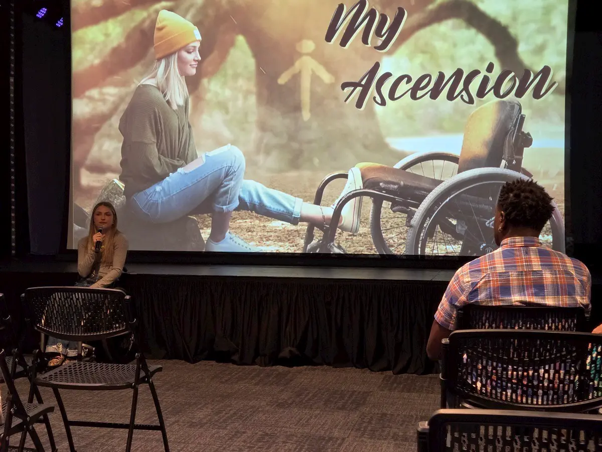 A woman speaks to a small audience in front of a large screen displaying an image of a woman sitting on the ground next to a wheelchair, with the words "My Ascension" written across the screen.