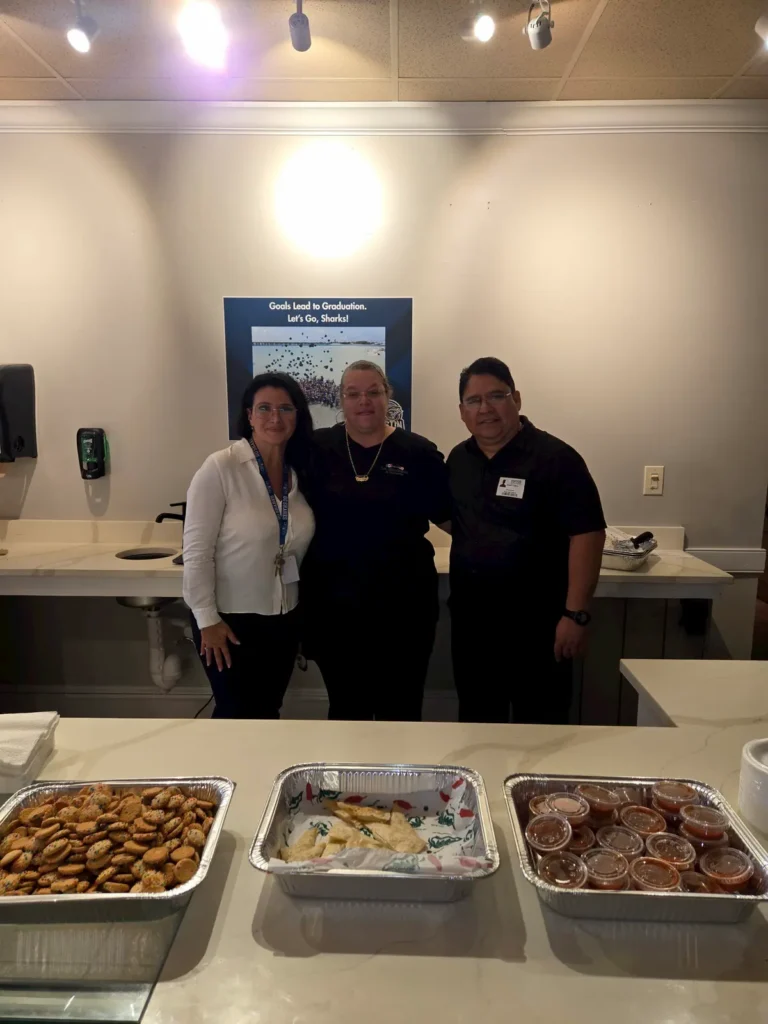 Three people stand behind a counter with trays of food, including desserts and sauces, in a well-lit room. They are smiling and posing for the photo. A sign and light fixtures are visible in the background.