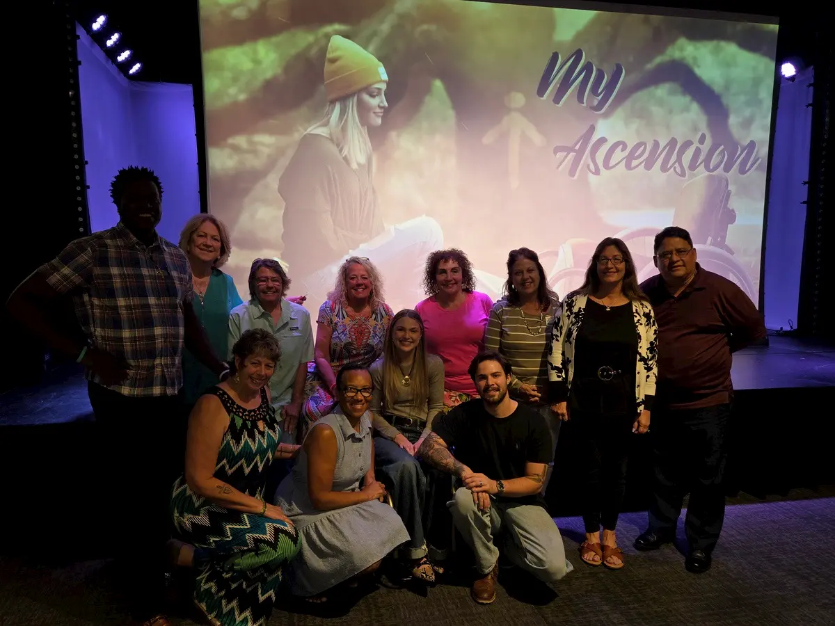 A group of 12 people pose together and smile in front of a projection screen displaying "My Ascension" and an image of a young woman in a beanie hat. The setting appears to be a theater or event space.