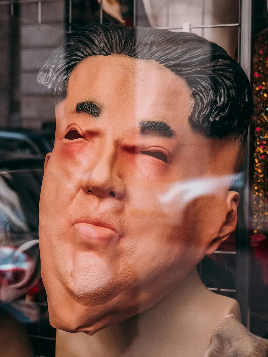 A rubber mask resembling a man's face with distinct features and slicked-back hair is displayed in a shop window, its expression subtly hinting at the characteristics of a bad leader, with reflections of the street visible on the glass.