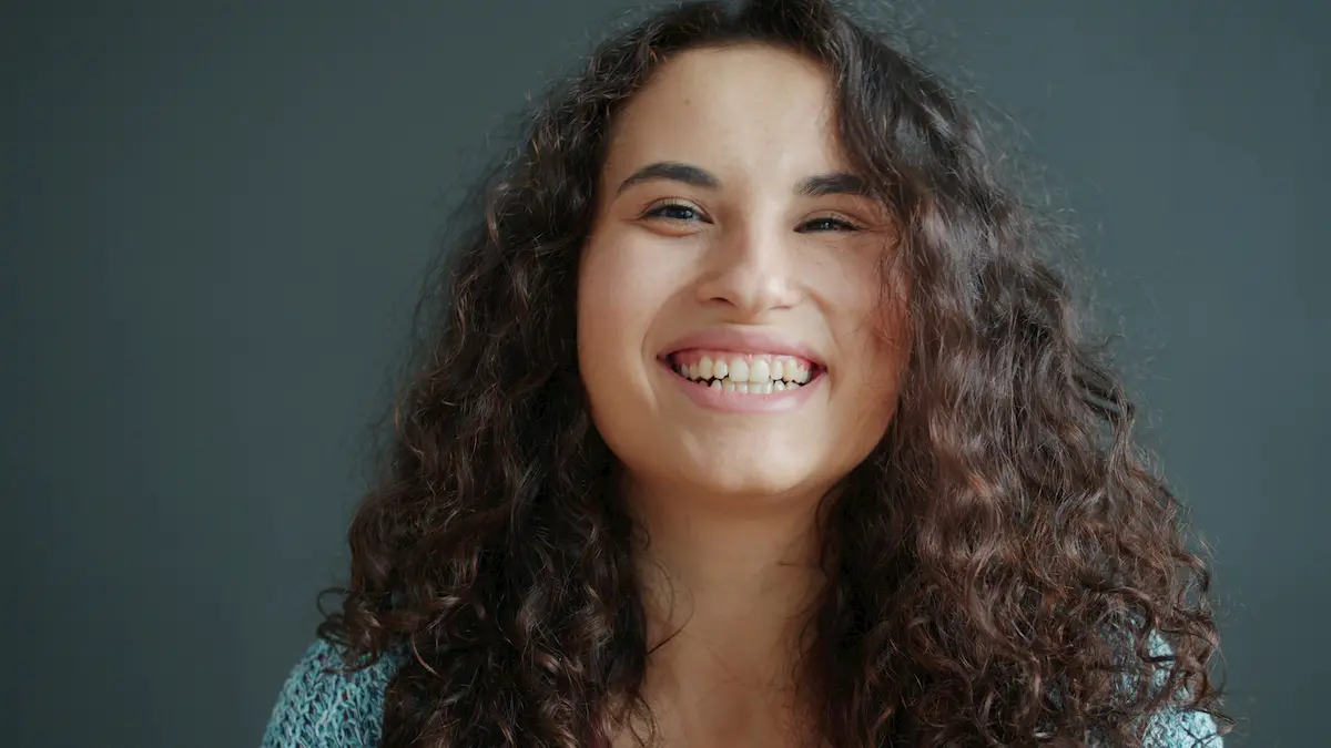 A young woman with long, curly brown hair smiles brightly at the camera against a plain, dark gray background. She is wearing a light blue top.