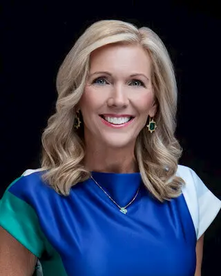 A smiling woman with blonde, wavy hair wearing a blue dress with green and white accents, gold earrings, and a necklace, posed against a black background.