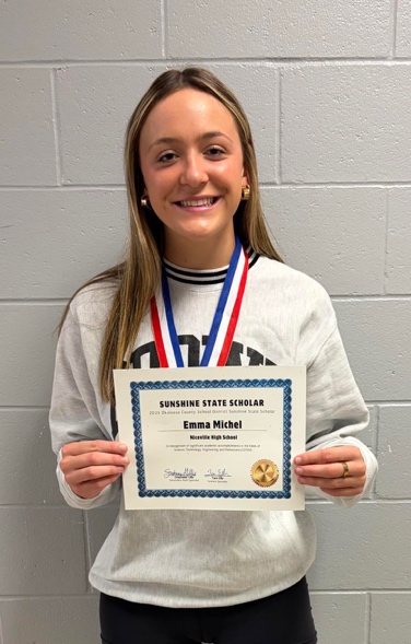 A smiling young woman wearing a white sweatshirt and two medals holds a “Sunshine State Scholar” certificate in front of a gray cinderblock wall.
