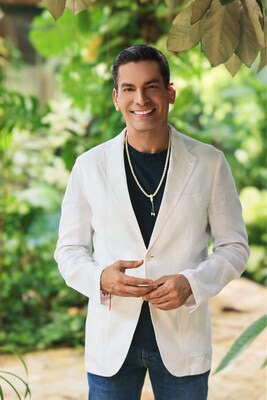 A man stands outdoors, smiling in a white blazer over a black shirt and blue jeans. He has short dark hair and is surrounded by lush green plants and sunlight, resembling Ismael Cala, the renowned international judge for Miss Universe 2025.